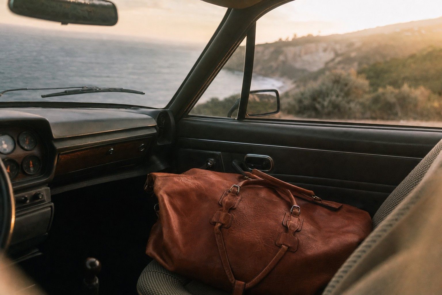 A leather duffle on the passenger seat of a classic car on a coast road
